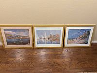 Three framed impressionist prints in gold frames depicting sailboats and a coastal scene, arranged side-by-side on hardwood floor.