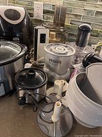 Photo shows air fryer, crockpot, Veggie Bullet food processor with attachments and white waffle maker on counter.
