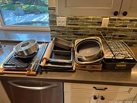 Wide view of roasting pan, racks, various baking pans, cookie sheets, cutting boards, and stove top grill pan arranged on kitchen counter.