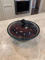 Photo showing a dark copper brown and teal ceramic bowl with a lid, highlighting the looped handle on top and glossy finish.