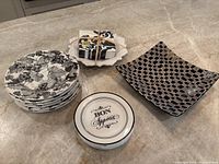 Overview of small black and white floral dessert plates stacked (8 total), round 'Bon Appetit' plates stacked (6 total), a black and white patterned square platter, and a bundle of cocktail napkins.