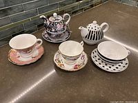 Tea sets displayed on countertop showing three sets of teapots with matching cups and saucers with floral and striped patterns.