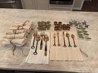 Full view of 12 place settings flatware laid out with burlap wrapped napkins and four styles of napkin rings on a marble countertop.