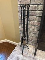 Black metal fireplace tool set with broom, poker, and shovel on a wrought iron stand with three legs, placed beside a brick fireplace.