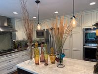 Overall kitchen countertop display of assorted colorful glass bottles, vases, and candleholders with dried ornamental grass held in some.