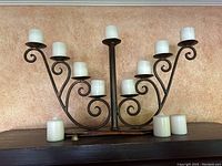 Wide view of brass candle holder with nine white candles mounted and three additional white candles placed in front of it on a dark surface against a beige textured background.