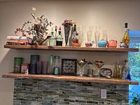 Two wooden shelves with various kitchen and decorative items including glassware, ceramic bowls, bottles, dried flower arrangements, stacked books, and artistic decorative pieces
