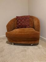 Front left angled view showing burnt orange velour fabric, rounded design, and brass rolling legs with a patterned pillow on seat.
