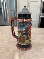 Side view of beer stein showing carved handle, lid, and painted figures of people sitting and drinking outdoors.