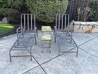 Two metal patio loungers with black rubber straps and reclining backs, alongside two wrought iron side tables with decorative tile tops, one chipped.