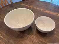 Photo shows two white ceramic bowls of different sizes on a wooden table, large deep bowl on the left and smaller bowl on the right.