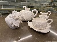 Side view of teapot, sugar bowl, creamer, and teacup with saucer showing floral embossing and glossy white finish.