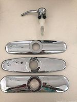 Three chrome escutcheon plates and one chrome-top soap dispenser pump laid out on surface