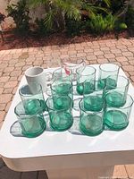 Tabletop view of 12 green glass tumblers with additional white mug, clear measuring cup and small clear cup