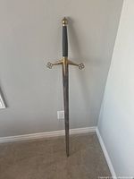 Full view of the antique long sword standing upright against a wall, showing the blade, crossguard, grip, and pommel.