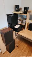 Large black subwoofer, two tall black speakers and amplifier unit arranged near a wooden shelf unit with visible wiring and power cord.
