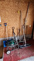 Full view of all garden tools arranged against a wooden wall, including hose reel, shovels, snow rakes, and other long-handled tools on concrete floor.