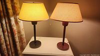 Both lamps on white table with shades on. Black lamp to left, burgundy lamp to right. Showing size and color difference.