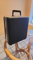 Full view of the closed black textured suitcase with metal edges on a wicker chair.