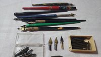 Full view of six varied straight dip pens with colored handles showing wear plus three separate nibs and a box of additional nibs
