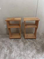 Two raw unfinished wood bedside tables side by side on carpet. Rectangular top, two open shelves below.