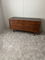 Front view of the wooden dresser showing six drawers and legs, placed against a plain wall on carpeted flooring.