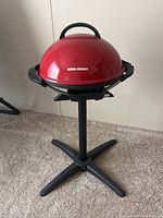Red dome-shaped George Foreman propane grill on black stand with tripod base, set on carpeted floor.