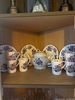 Photo showing 6 floral pattern cups, 6 saucers, a teapot with lid all with floral pattern and gold trim on wooden shelf