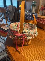 View of rectangular woven wood basket with wooden handle and floral fabric liner tied by pink ribbon on wooden table.
