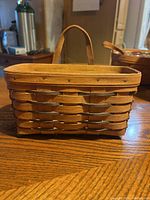 Small rectangular Longaberger basket with single handle on a wooden table under indoor lighting, showing overall front and side view.