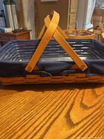 Side view of rectangular woven Longaberger basket with wooden handles resting on table, showing weaving pattern and fabric liner edge.