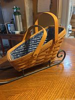 Side and angled views of the Longaberger woven basket lined with blue floral fabric, sitting in black metal sleigh holder stand on wooden table.