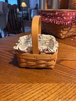 Small woven wooden Longaberger basket with floral cloth liner on wooden table, showing full basket shape and handle.