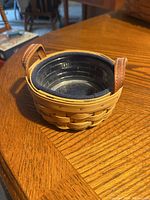 Side view showing woven basket with black plastic liner inside and two brown leather handles.