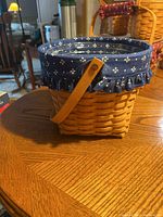 Longaberger rectangular basket with wooden handle and blue floral fabric liner with ruffle on wooden table.