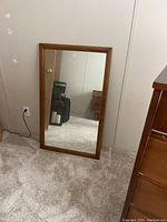 Front view of rectangular mirror with brown wood frame leaning against wall on carpeted floor showing reflective glass surface.