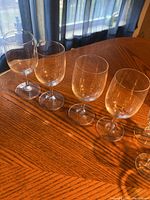 Four clear wine glasses with rounded bowls, arranged on wooden table near window showing natural light and curtains in background.