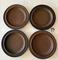 Four round brown and dark brown ceramic plates arranged flat showing surface and rim speckled texture.