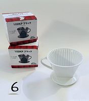 Two boxed Kalita 102KP filter holders in black plastic next to a white Melitta ceramic filter holder with handle and ribbed interior.