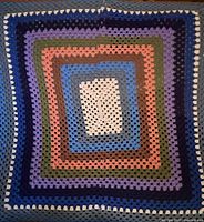 Full view of the crocheted wool blanket showing square pattern with multiple concentric color bands including white, grey, blue, pink, olive green, purple and dark blue.