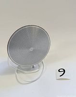 Front view of circular speaker with white perforated grill and wood finish stand.