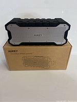 Front view of Aukey SoundTank wireless speaker on original cardboard box showing silver grille and black rugged edges