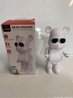 Photo showing the E-mrge Bear Speaker in its box, white bear figure with black sunglasses beside it, box featuring product image and brand name.