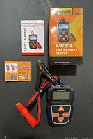 Photo shows the KONNWEI KW208 car battery tester device with LCD screen, associated test cables with clamps, the user manual, an instruction or accessory card, and the product box standing upright.