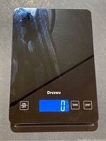 Full view of the black Drcowu digital food scale showing some surface scratches and smudges with the LCD turned on displaying zero weight.