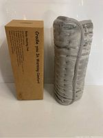 Rolled gray heating pad next to its cardboard box labeled 'Cradle You in Warming Comfort'. The box mentions body heating pad and warming comfort features.