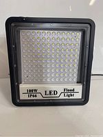 Front view of black framed LED flood light showing LED array and label indicating 100W and IP66 rating.