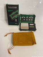 Fuzion FU-200 professional digital scale in green and black with weighing platform, digital display, control buttons, original box, and yellow drawstring pouch.