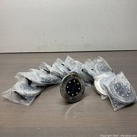 12 solar LED ground lights laid out on a flat surface, individually bubble wrapped except one light visible showing the stainless steel frame and solar panel with LEDs.