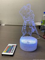 3D LED illusion lamp of anime character with white textured base and multicolor remote on table, powered by cable.